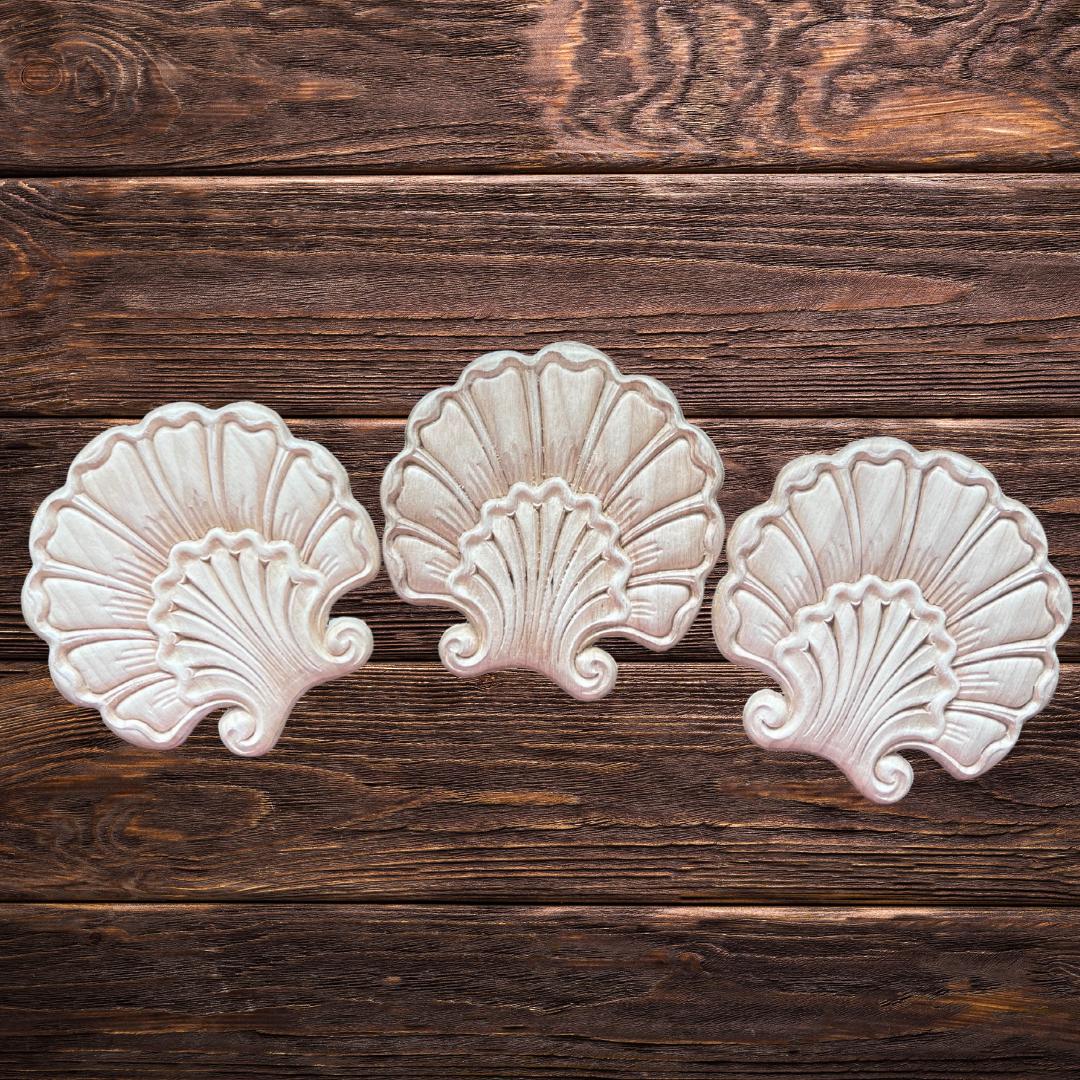 Three decorative, wooden shell-shaped appliques are arranged in a row on a rustic wooden surface with visible grain patterns.