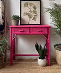 A vibrant pink console table stands against a white wall, topped with a botanical framed print. Potted plants of varying sizes and textures surround the table. The setting is styled in a cozy, bohemian decor with a woven rug beneath.