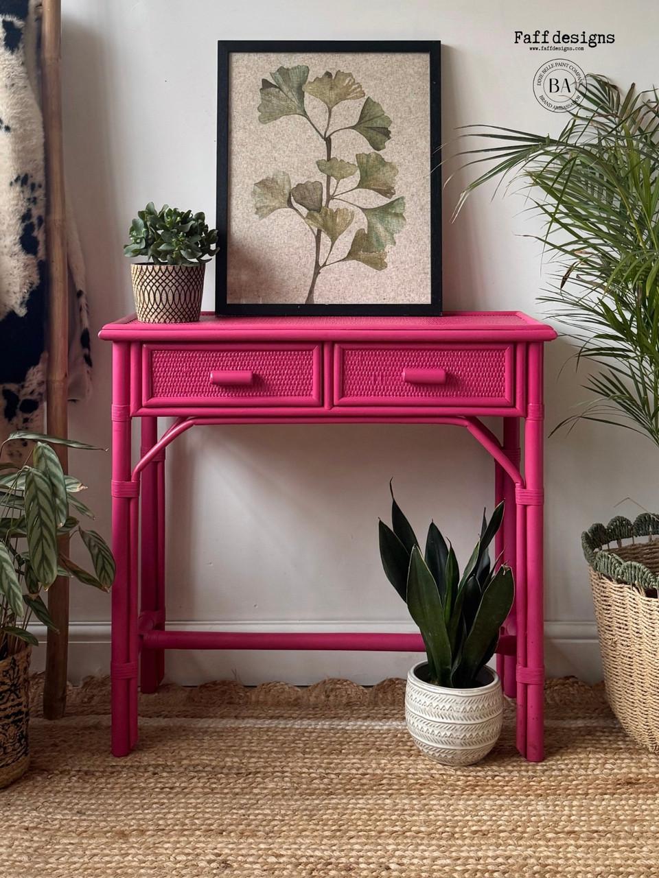A vibrant pink console table stands against a white wall, topped with a botanical framed print. Potted plants of varying sizes and textures surround the table. The setting is styled in a cozy, bohemian decor with a woven rug beneath.