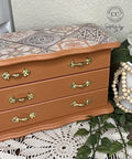 A small, decorative chest with three drawers featuring gold handles and a patterned top sits on a white surface next to a potted plant and a white beaded vase holding white flowers.