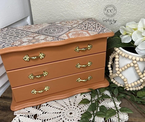 A small, decorative chest with three drawers featuring gold handles and a patterned top sits on a white surface next to a potted plant and a white beaded vase holding white flowers.