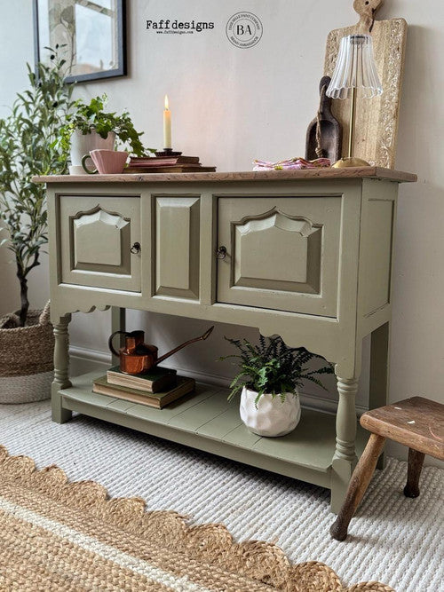 A small green sideboard with two paneled doors stands against a wall, topped with decor items, a lit candle, and books. A plant and books sit on the lower shelf; a woven rug and basket are nearby.