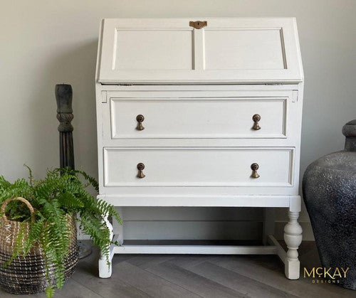 A white vintage writing desk with two drawers, brass handles, and slanted top stands against a light wall. A fern in a woven basket and a decorative sculpture are on the floor beside it. The McKAY Designs watermark is in the corner.