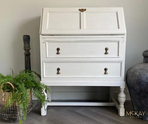 A white vintage writing desk with two drawers, brass handles, and slanted top stands against a light wall. A fern in a woven basket and a decorative sculpture are on the floor beside it. The McKAY Designs watermark is in the corner.