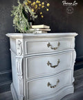A vintage-style gray dresser with ornate brass handles stands against a dark patterned wall. A vase with yellow and green flowers, along with stacked books, adorns the top. The floor is a dark wood. The photo and piece is by Theresa Lee.