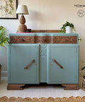 A vintage sideboard with ornate wood carvings is painted in light blue. It features two large cabinet doors with wooden handles. Potted plants and a table lamp with a woven base are placed on and around it. The setting includes a woven rug.