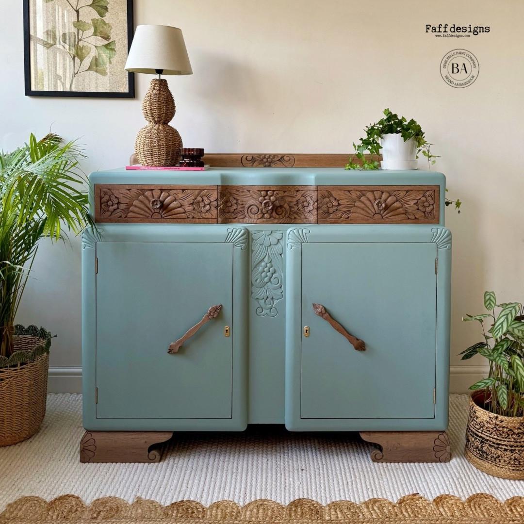 A vintage sideboard with ornate wood carvings is painted in light blue. It features two large cabinet doors with wooden handles. Potted plants and a table lamp with a woven base are placed on and around it. The setting includes a woven rug.