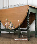 Wooden table with a foldable top, decorated with floral designs and labeled "Whiskey Barrel." Finished with a water-based stain, it boasts dark green, twisted legs. Text at the top reads "Gracie's House Painted Furniture," set against a white paneled wall.