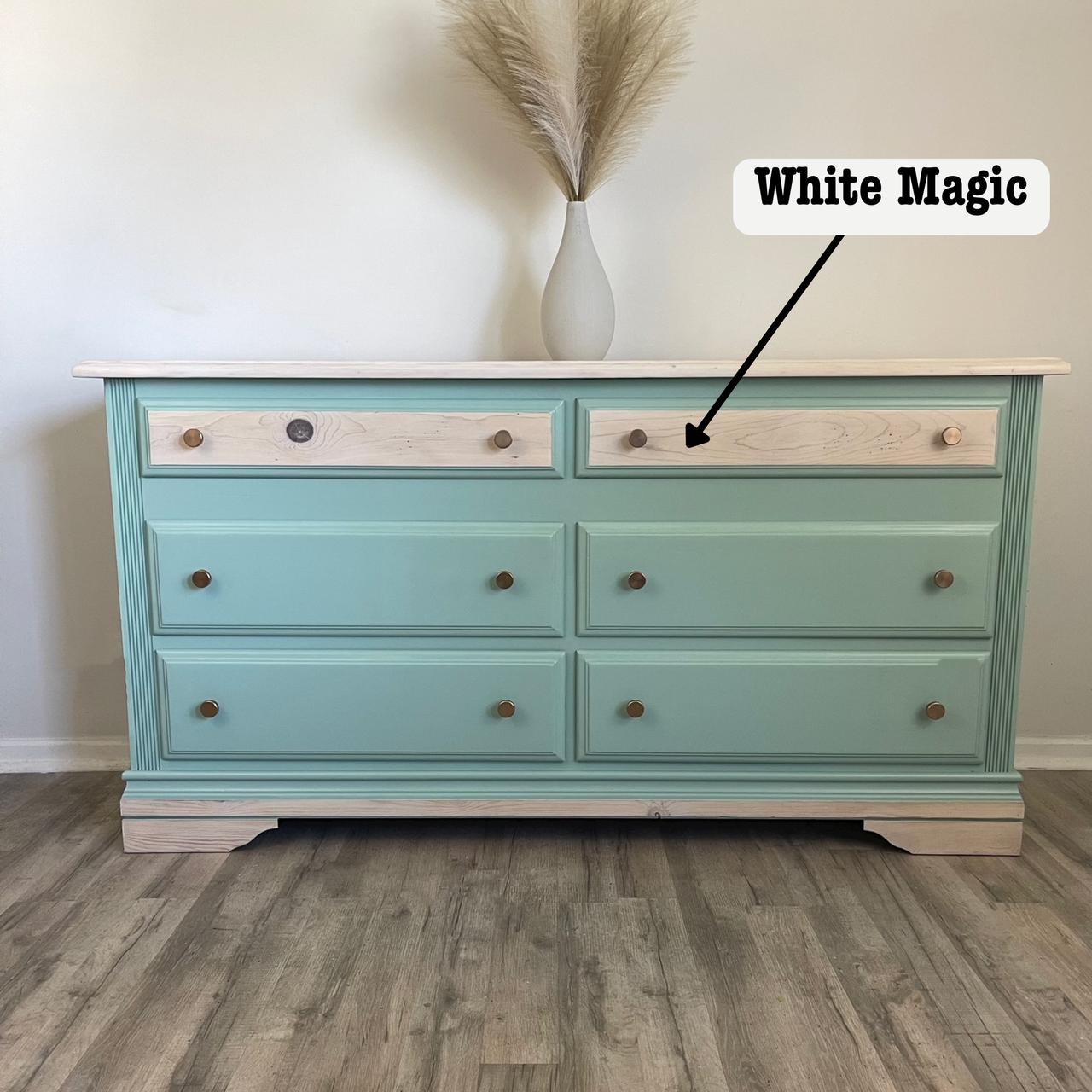 A dresser with six teal drawers and two light wood drawers on top. There's a vase with pampas grass on top. An arrow points to one of the light wood drawers labeled "White Magic." The floor is wooden.