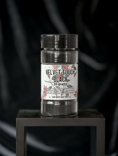 A container labeled "Velvet Touch Black, 30 grams" sits on a black square stand against a dark, velvety background. The label includes floral designs and a warning: "Do not get wet."