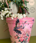 A pink Flower pot. Versailles Garden Transfer is seen applied around the pot. A blurry Wreath in the back ground above the pot with white flowers on the rim of the flower pot.
