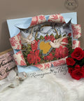 A decorative frame featuring two doves surrounding a heart and floral design, embellished with red and blue patterned ribbon. Nearby are red roses, two ceramic doves, and a wooden sign reading "Forever be my Always," all on a soft white surface.