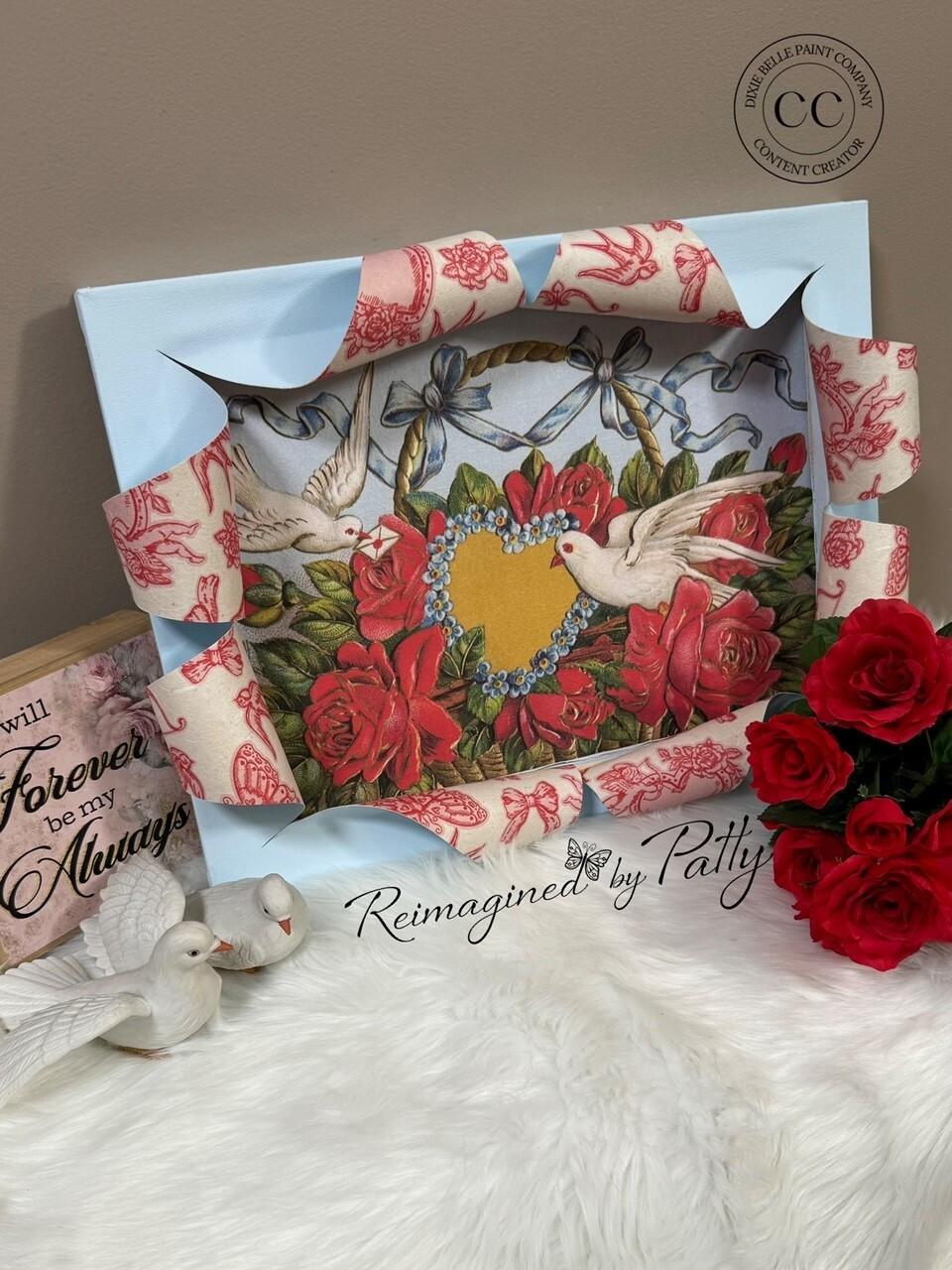 A decorative frame featuring two doves surrounding a heart and floral design, embellished with red and blue patterned ribbon. Nearby are red roses, two ceramic doves, and a wooden sign reading "Forever be my Always," all on a soft white surface.
