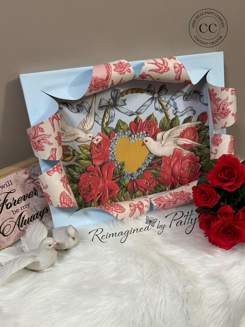 A decorative frame featuring two doves surrounding a heart and floral design, embellished with red and blue patterned ribbon. Nearby are red roses, two ceramic doves, and a wooden sign reading "Forever be my Always," all on a soft white surface.