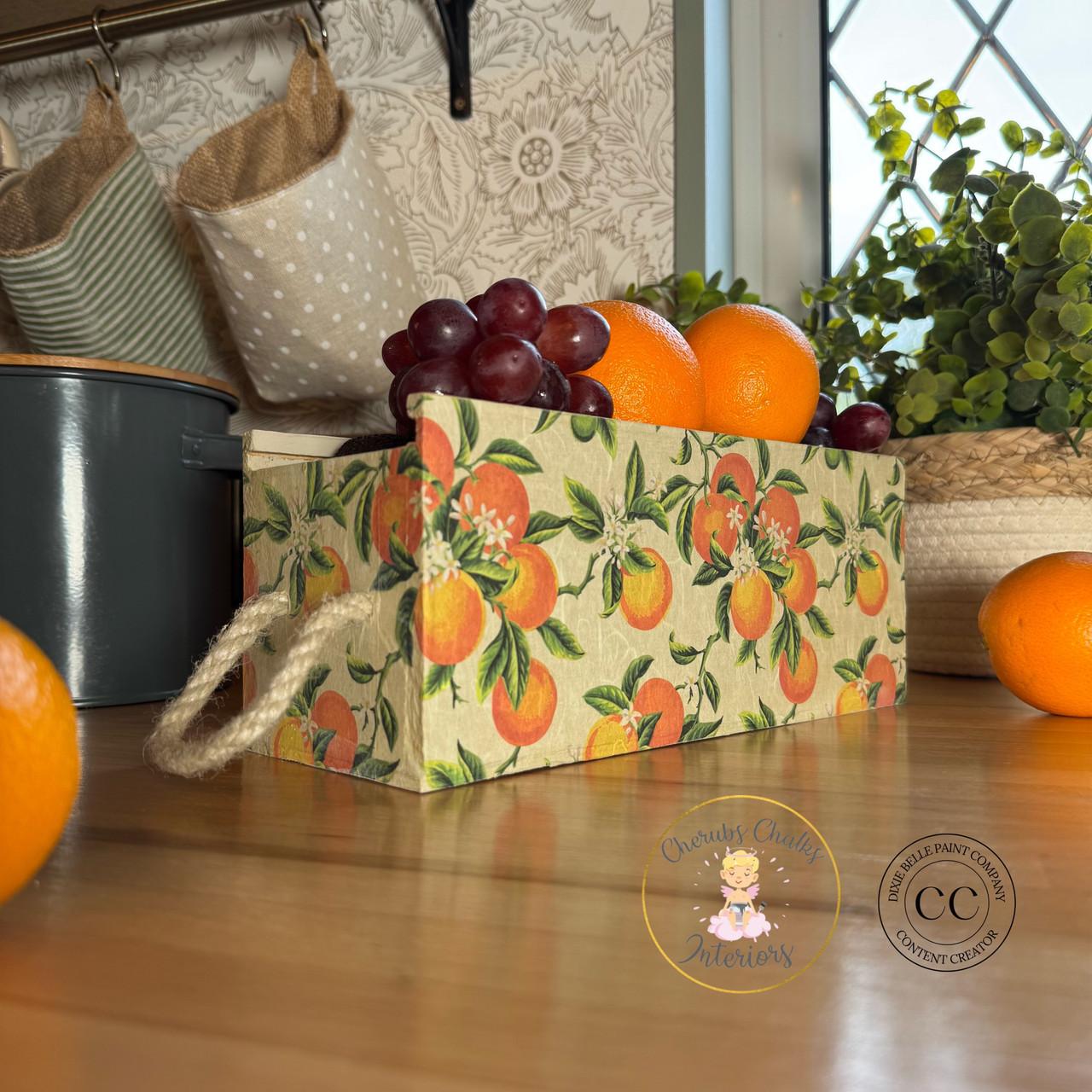 A decorative box with orange and green floral patterns with oranges is filled with grapes and oranges, placed on a wooden countertop. A rope handle is on the side of the box. Kitchen items and a window are in the background.