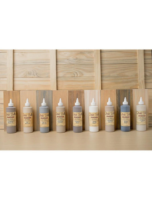 Nine bottles of Dixie Belle VooDoo Stains in varying shades are lined up on a table. The background features wooden planks in different natural tones, complementing the stains.