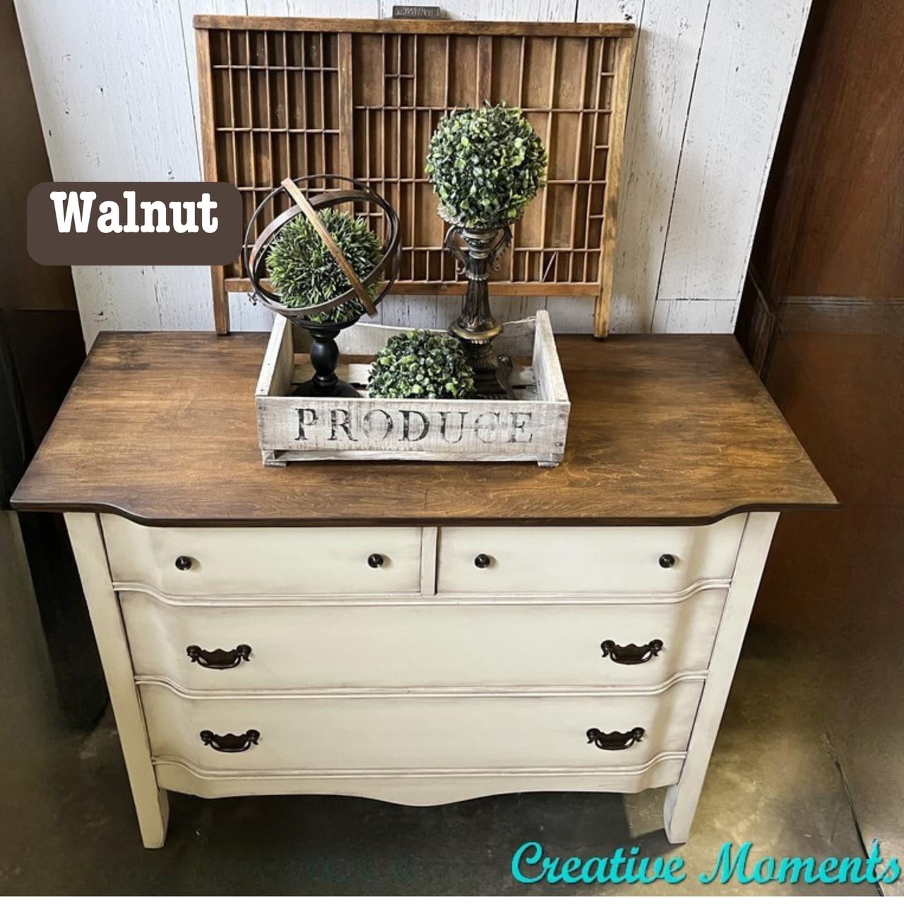 A vintage dresser with a dark walnut top and cream-colored body. It has ornate drawer handles. On top, there's a decorative tray holding two small bush sculptures and a circular metal decor piece. A "Creative Moments" watermark is at the bottom.
