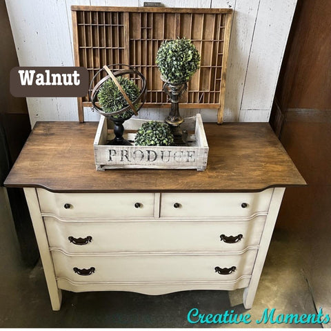A vintage dresser with a dark walnut top and cream-colored body. It has ornate drawer handles. On top, there's a decorative tray holding two small bush sculptures and a circular metal decor piece. A "Creative Moments" watermark is at the bottom.