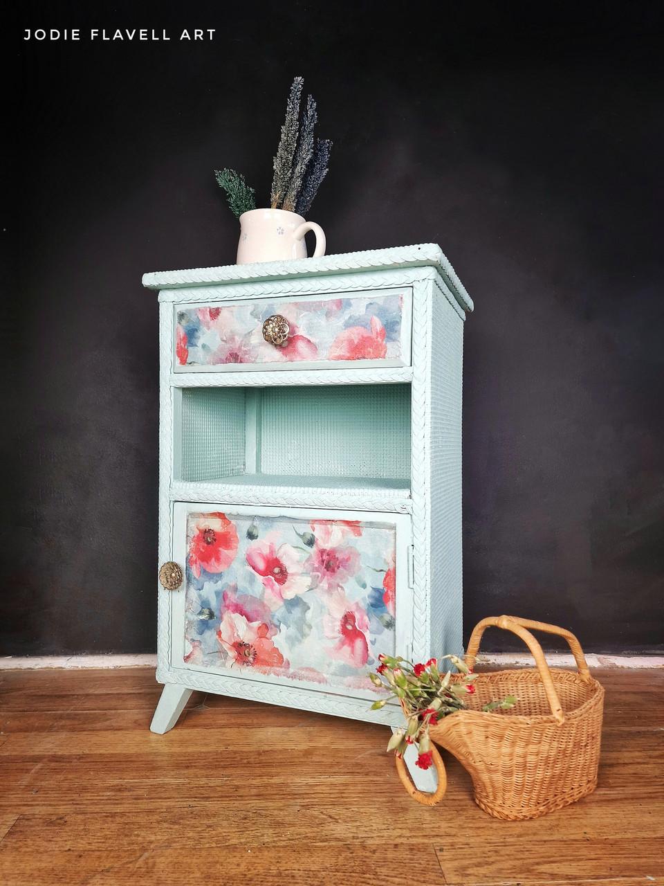 A small painted cabinet with floral designs stands on a wooden floor against a dark wall. It has one drawer, a shelf, and a door. A bouquet in a white mug is on top, and a wicker basket with flowers is beside it. "Jodie Flavell Art" is written above.