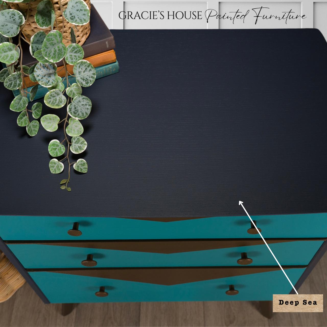 Overhead view of a dark blue, three-drawer chest labeled "Deep Sea," finished with Deep Sea Silk All-in-One paint. The teal drawers feature dark knobs. A potted plant and books rest atop. Text at the top reads "Gracie's House Painted Furniture.
