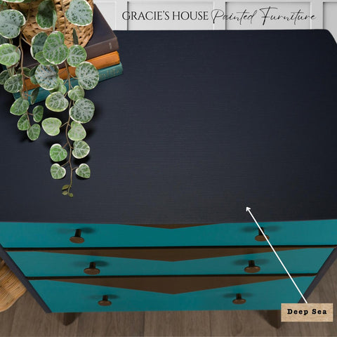 Overhead view of a dark blue, three-drawer chest labeled "Deep Sea," finished with Deep Sea Silk All-in-One paint. The teal drawers feature dark knobs. A potted plant and books rest atop. Text at the top reads "Gracie's House Painted Furniture.