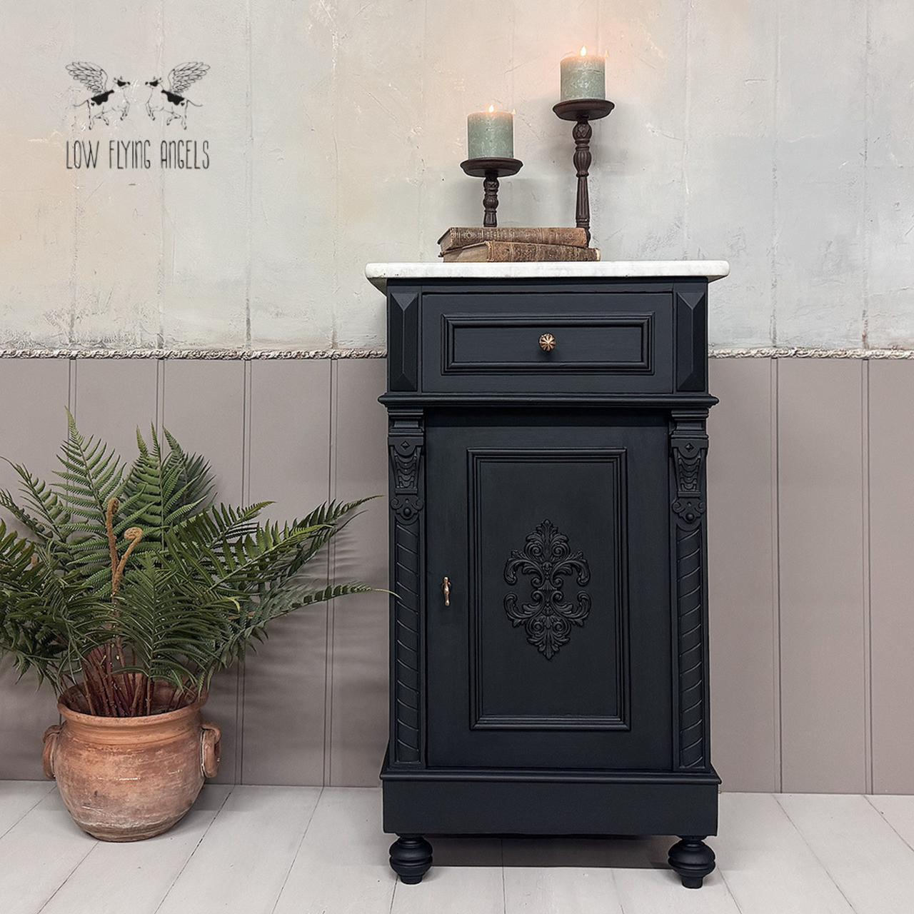 A black vintage cabinet with ornate detailing is placed against a two-tone wall. Two lit candles on holders sit on top. An orange pot with green plants is on the left. The background has a "Low Flying Angels" design.