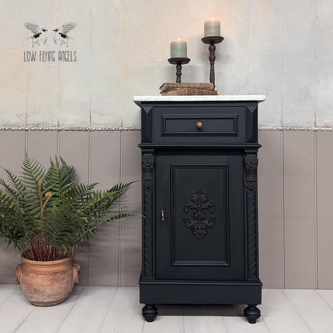 A black vintage cabinet with ornate detailing is placed against a two-tone wall. Two lit candles on holders sit on top. An orange pot with green plants is on the left. The background has a "Low Flying Angels" design.