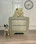 A mute green nightstand with Wedding Lace Transfer seen applied to the fronts of the drawer has gold hardware. Staged next to a wooden black and white dog statue and a short vase with large white flowers cover majority of it on top of the nightstand.