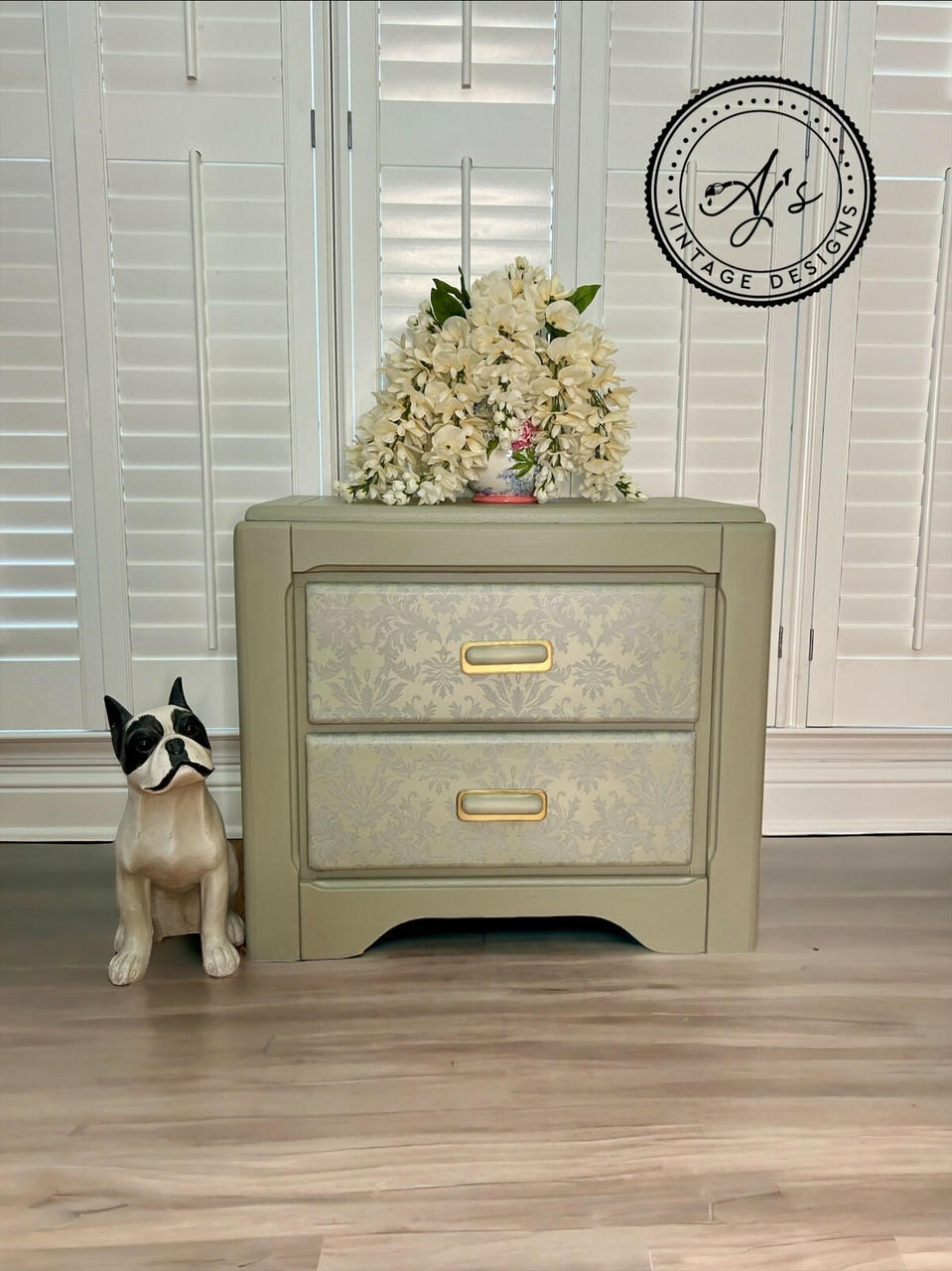 A mute green nightstand with Wedding Lace Transfer seen applied to the fronts of the drawer has gold hardware. Staged next to a wooden black and white dog statue and a short vase with large white flowers cover majority of it on top of the nightstand.