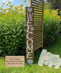 A tall wooden sign with "WELCOME" painted vertically in white letters stands in a garden with yellow flowers. Floral designs decorate the sign. A label reads: "Tea Party Bouquet Transfer applied on top."