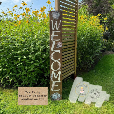 A tall wooden sign with "WELCOME" painted vertically in white letters stands in a garden with yellow flowers. Floral designs decorate the sign. A label reads: "Tea Party Bouquet Transfer applied on top."