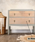 A light wooden dresser with antique handles, labeled "Au Naturel," stands on a wooden floor next to a woven basket with a white fringed cloth. The background has a soft, neutral-toned wall.
