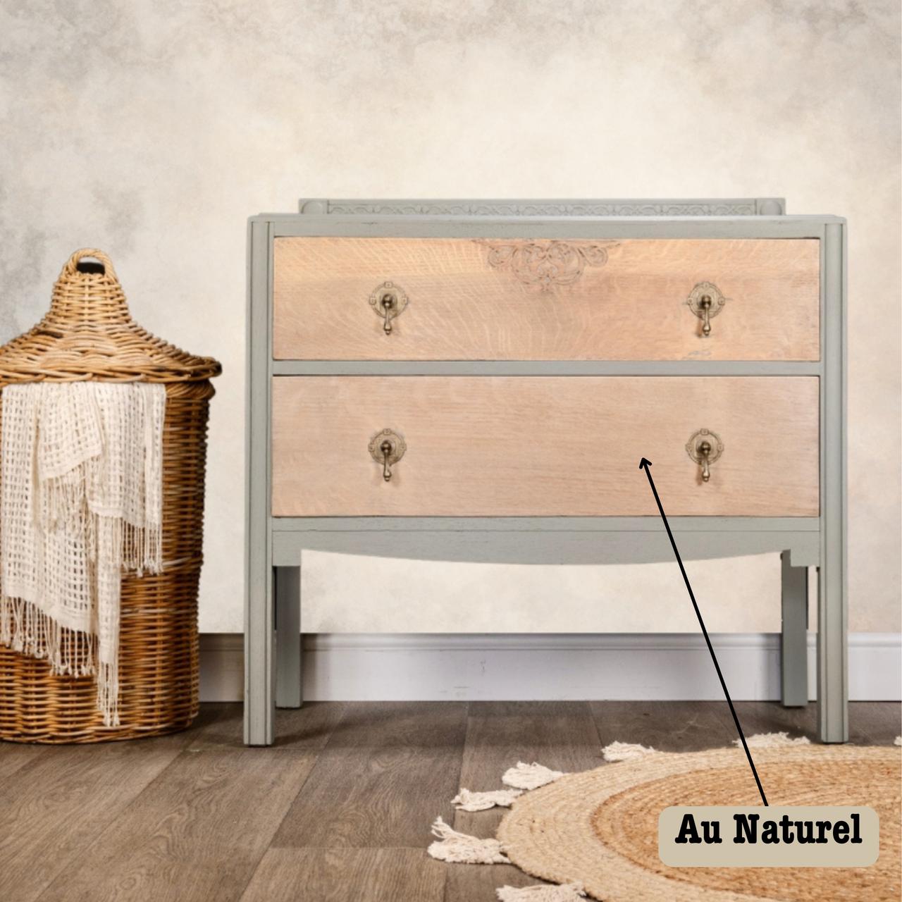 A light wooden dresser with antique handles, labeled "Au Naturel," stands on a wooden floor next to a woven basket with a white fringed cloth. The background has a soft, neutral-toned wall.