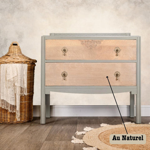 A light wooden dresser with antique handles, labeled "Au Naturel," stands on a wooden floor next to a woven basket with a white fringed cloth. The background has a soft, neutral-toned wall.
