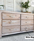 A rustic wooden dresser with six drawers and metal handles. A potted plant and decor sit on top. A label reads "White Magic" pointing to the dresser. The background features white paneled walls and a patterned rug.