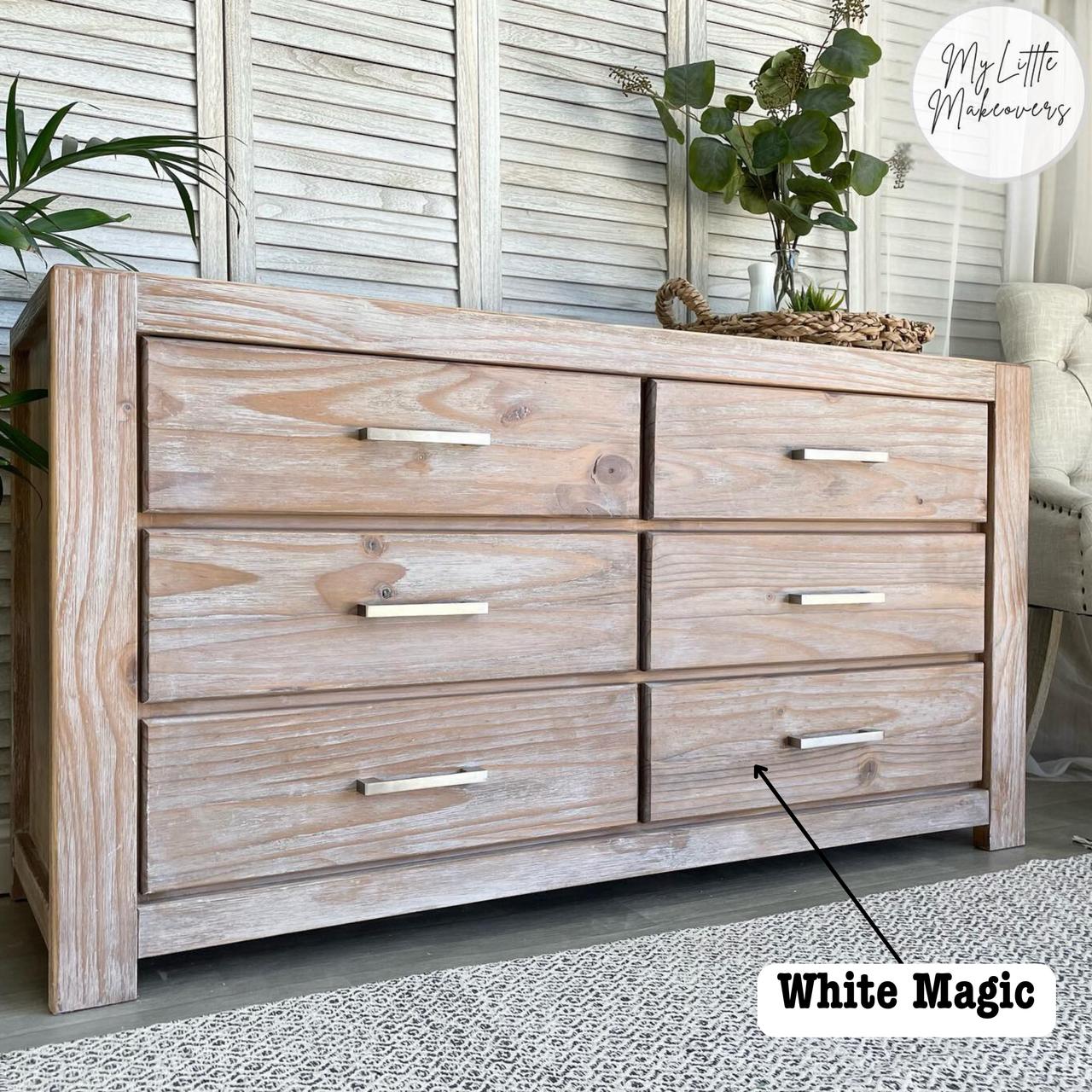A rustic wooden dresser with six drawers and metal handles. A potted plant and decor sit on top. A label reads "White Magic" pointing to the dresser. The background features white paneled walls and a patterned rug.