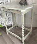 A vintage side table with twisted legs, painted in pale green and white with a floral design on top, stands on a wooden floor beside a decorative screen, a basket, and a fern plant.
