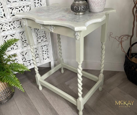 A vintage side table with twisted legs, painted in pale green and white with a floral design on top, stands on a wooden floor beside a decorative screen, a basket, and a fern plant.