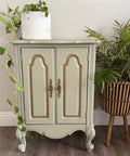 A small, light green vintage cabinet with gold accents stands against a wall. A white pot with a trailing plant sits on top, and a leafy potted plant in a striped planter is placed to the right.