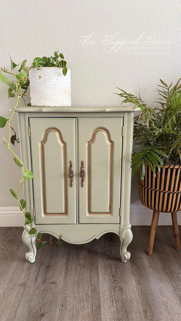 A small, light green vintage cabinet with gold accents stands against a wall. A white pot with a trailing plant sits on top, and a leafy potted plant in a striped planter is placed to the right.