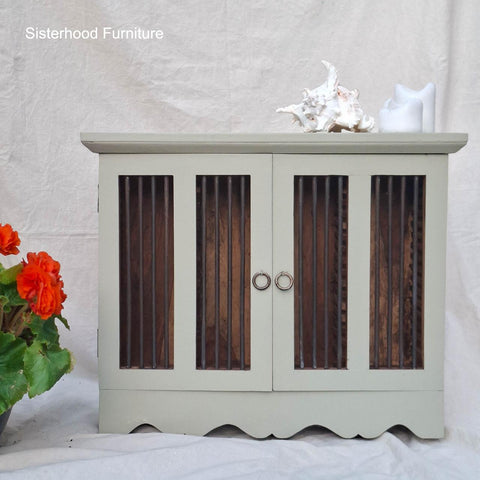 A small, light green cabinet with glass-paneled doors and metal handles, decorated with white seashells and a candle on top. A red flower plant is placed to the left of the cabinet.