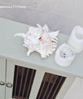 A large white seashell and two melted white candles rest on top of a pale green cabinet with glass doors. The text "Sisterhood Furniture" appears in the upper left corner.