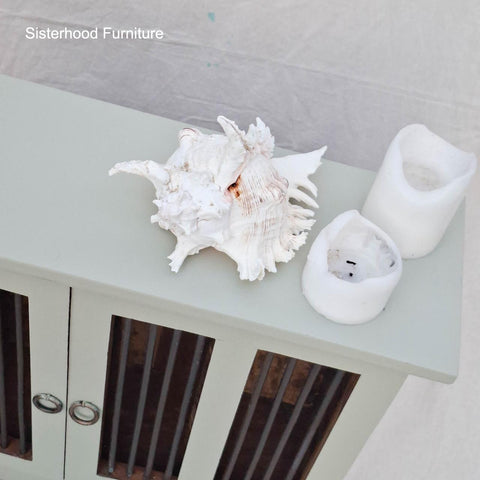 A large white seashell and two melted white candles rest on top of a pale green cabinet with glass doors. The text "Sisterhood Furniture" appears in the upper left corner.