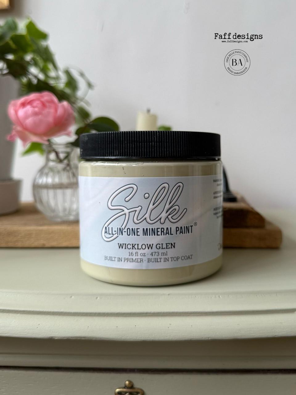A jar of Silk All-in-One Mineral Paint, labeled "Wicklow Glen," sits on a light-colored table. In the background, there is a pink rose in a vase and blurred decorative items.