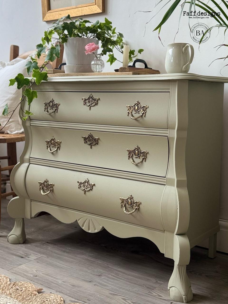 A vintage-style sage green dresser with ornate gold handles stands against a white wall, decorated with potted plants, a mug, a tray, and a picture frame on top. The dresser features curved legs and intricate detailing.