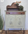 A vintage wooden sideboard with a decorative hutch, painted light green with dark wood accents. It has ornate details, floral and plant decor on top, and stands on turned legs against a white brick wall.