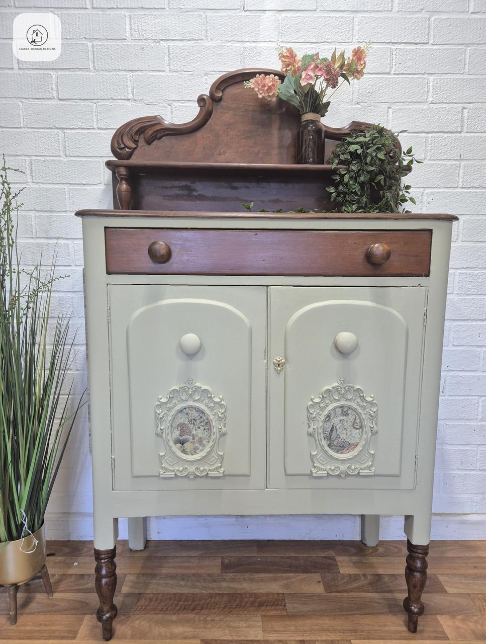 A vintage wooden sideboard with a decorative hutch, painted light green with dark wood accents. It has ornate details, floral and plant decor on top, and stands on turned legs against a white brick wall.