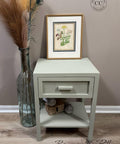 A light green nightstand with a single drawer and shelf stands against a beige wall. On top is a framed botanical print, and beside it is a tall glass vase with dried grasses. The floor is light wood.