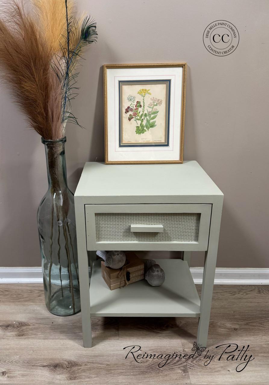 A light green nightstand with a single drawer and shelf stands against a beige wall. On top is a framed botanical print, and beside it is a tall glass vase with dried grasses. The floor is light wood.