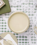 A jar filled with pale green paint sits on a lace and green checkered tablecloth, surrounded by a palette knife, a green book, and part of a teacup.